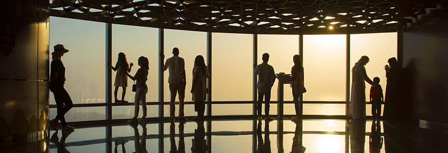 Silhouettes of people standing in an observation deck with floor-to-ceiling windows overlooking a cityscape at sunset